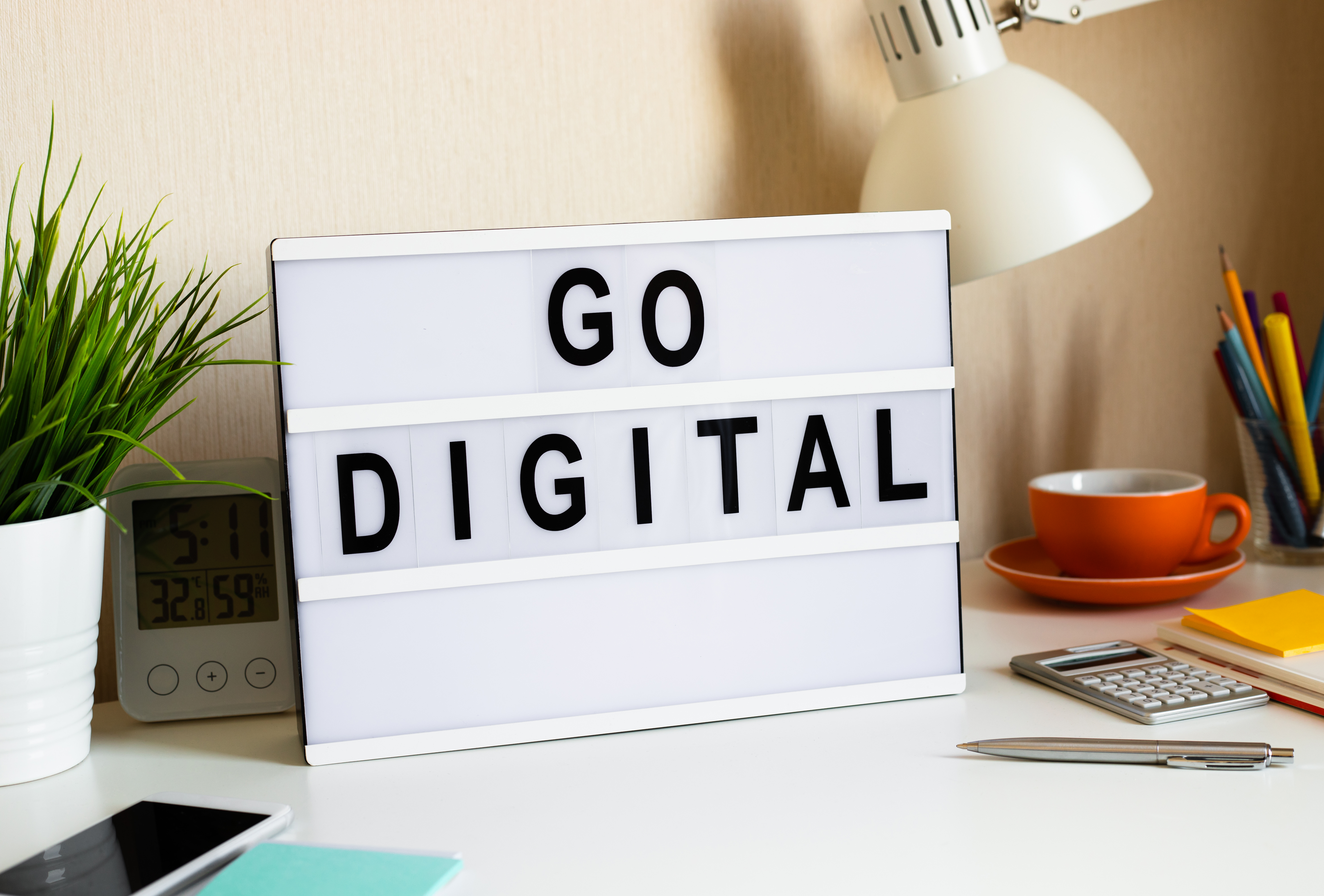 A modern desk setup featuring a lightbox sign that reads "Go Digital," surrounded by a green plant, office supplies, and a digital clock. Perfect imagery to emphasize the transition to digital flight scheduling solutions and the benefits of streamlining operations in a flight school or aviation business.