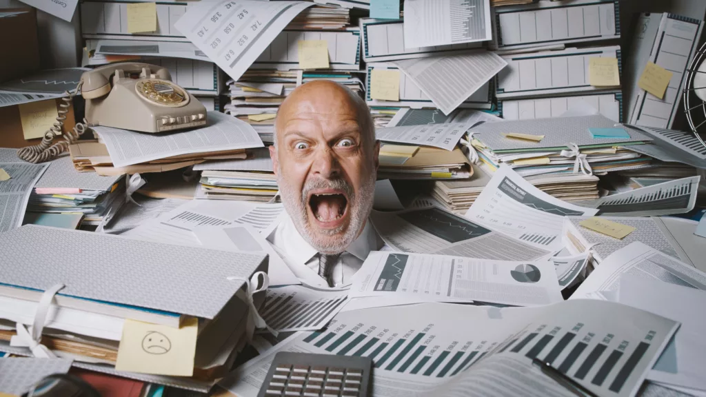 Stressed bald man in a white shirt and tie, screaming while buried under a chaotic pile of paperwork, folders, and financial reports. An old rotary phone sits on top of the clutter. The scene represents scheduling chaos, emphasizing the need for a modern solution like a flight schedule maker like PreFlight to eliminate overwhelming paperwork and confusion.