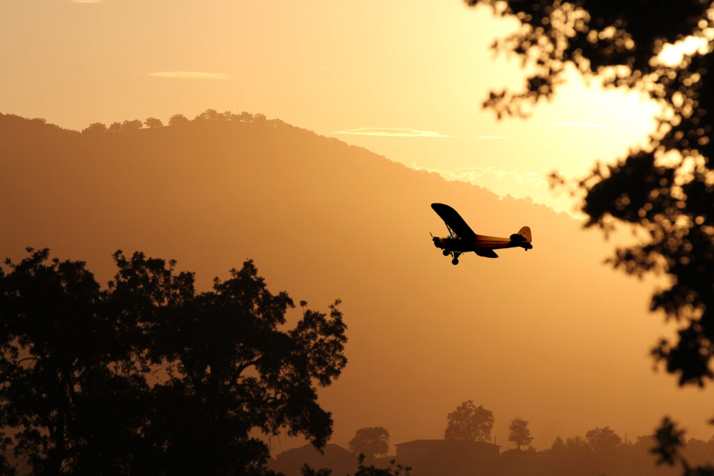 Small single-engine aircraft flying at sunset over a landscape of rolling hills and silhouetted trees, representing the freedom and adventure of aviation. Perfect for illustrating seamless aircraft booking for Intro Flights.